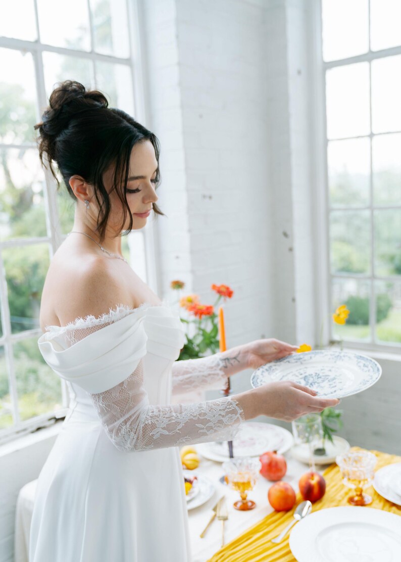 May include: A white off-the-shoulder dress with lace sleeves. A woman is holding a white plate with a blue floral design. The table is set with plates, glasses, fruit, and flowers. The scene is set in front of a large window.