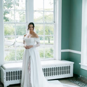 May include: A bride in a white off-the-shoulder wedding dress with a long train and a high slit. The dress features lace sleeves and a fitted bodice. The bride is standing in front of a large window with a white bench.