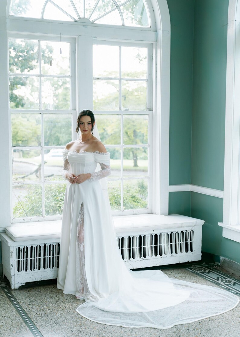 May include: A bride in a white off-the-shoulder wedding dress with a long train and a high slit. The dress features lace sleeves and a fitted bodice. The bride is standing in front of a large window with a white bench.