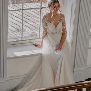 May include: A bride wearing a white lace wedding dress with a tulle skirt. She is standing in front of a window with natural light streaming in.