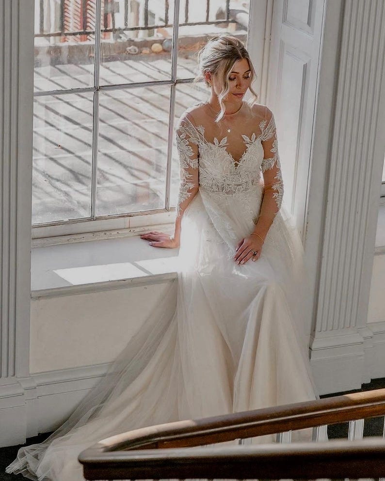 May include: A bride wearing a white lace wedding dress with a tulle skirt. She is standing in front of a window with natural light streaming in.
