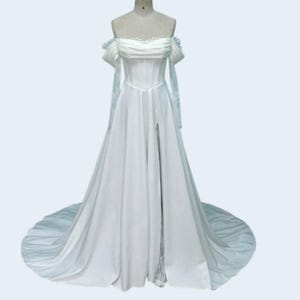 May include: A white off-the-shoulder wedding dress with a corset bodice and a high slit. The dress features delicate lace sleeves and a flowing skirt that extends into a train. The dress is displayed on a mannequin.