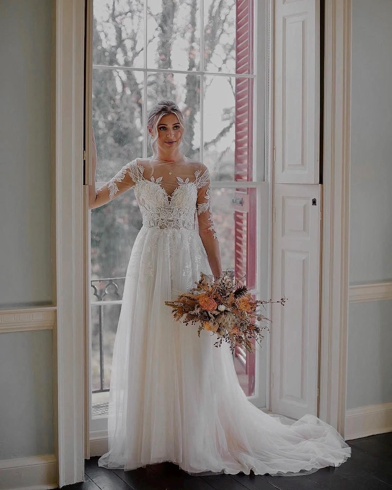 May include: A bride in a white lace wedding dress with long sleeves and a flowing skirt. She is holding a bouquet of dried flowers in shades of orange and brown. The bride is standing in front of a window with a view of trees.