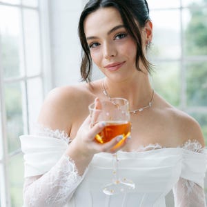 May include: A woman in a white off-the-shoulder dress holds a glass of amber liquid. The dress features lace sleeves and a corset-style bodice. She wears a delicate necklace and earrings. The background includes a window with natural light.