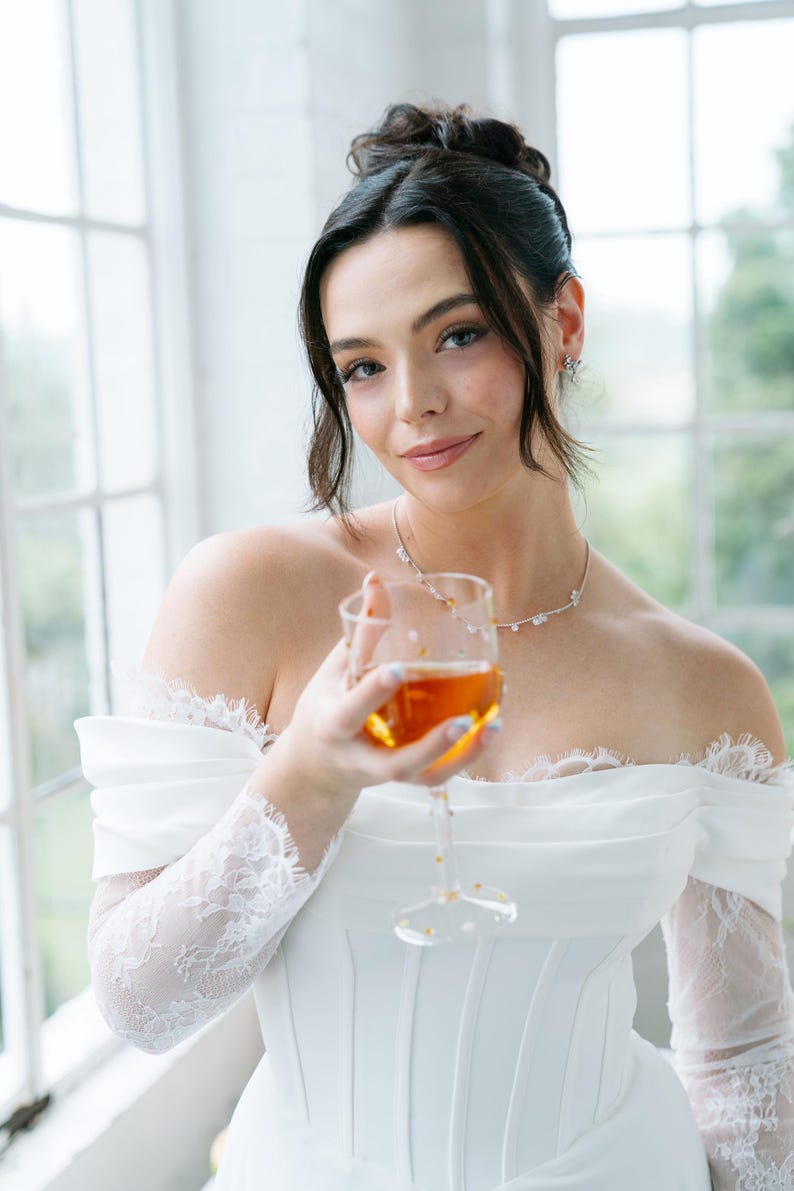 May include: A woman in a white off-the-shoulder dress holds a glass of amber liquid. The dress features lace sleeves and a corset-style bodice. She wears a delicate necklace and earrings. The background includes a window with natural light.