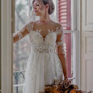 May include: A bride wearing a white lace wedding dress with long sleeves and a flowing skirt. She is holding a bouquet of dried flowers in shades of brown and orange.