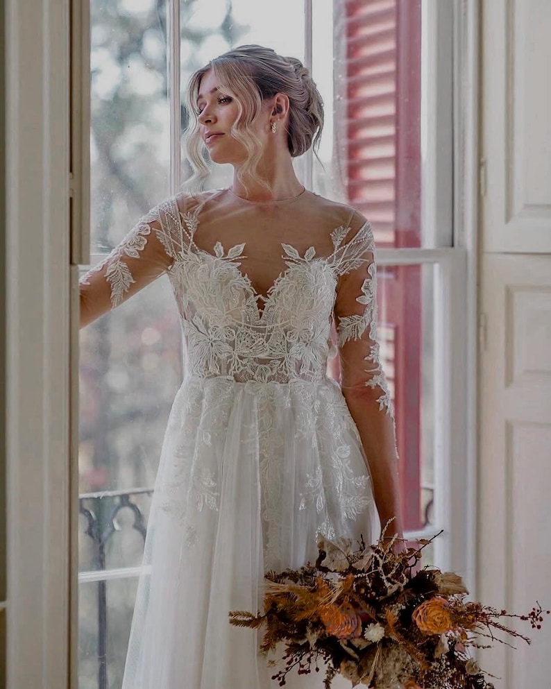 May include: A bride wearing a white lace wedding dress with long sleeves and a flowing skirt. She is holding a bouquet of dried flowers in shades of brown and orange.