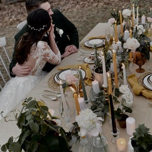 May include: A bride and groom sit at a table decorated with white and yellow candles, greenery, and white flowers. The table is set with white plates, gold chargers, and yellow napkins.