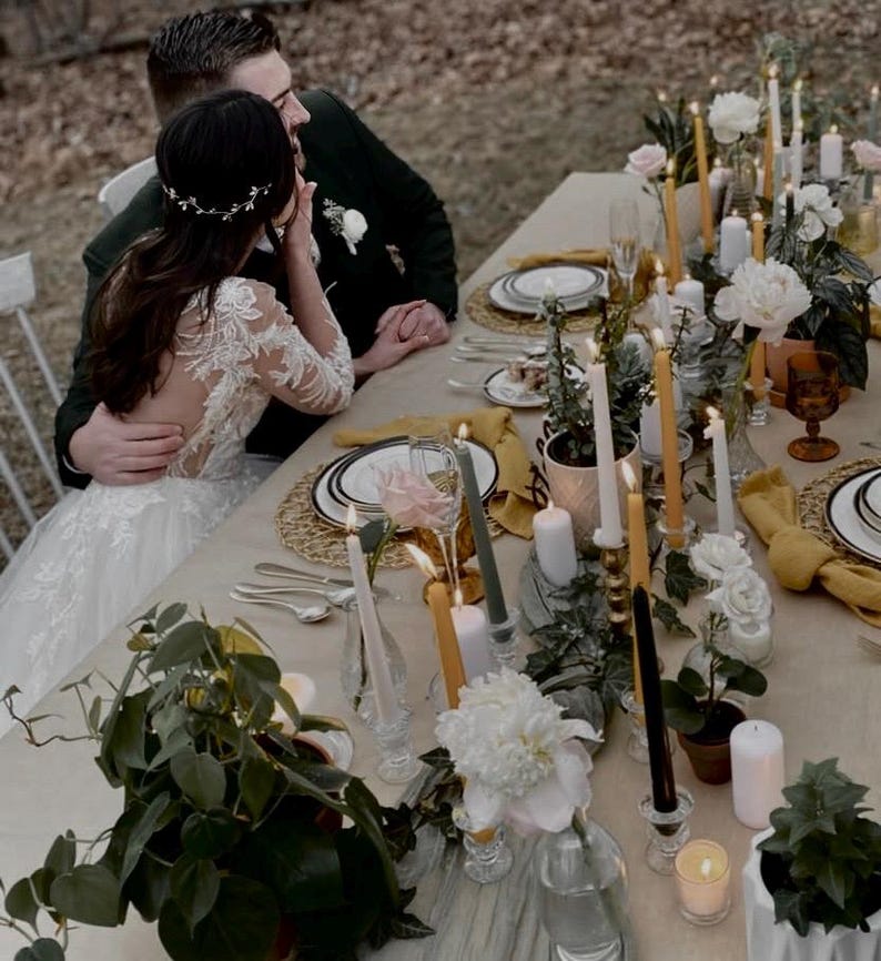 May include: A bride and groom sit at a table decorated with white and yellow candles, greenery, and white flowers. The table is set with white plates, gold chargers, and yellow napkins.