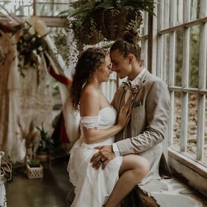 May include: A couple embraces in a sunlit room, the woman in a white lace wedding dress and the man in a tan suit. The scene is set near a window with hanging plants, suggesting a wedding or engagement photoshoot.