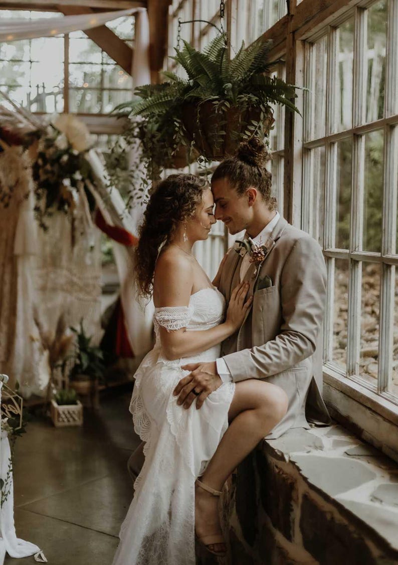 May include: A couple embraces in a sunlit room, the woman in a white lace wedding dress and the man in a tan suit. The scene is set near a window with hanging plants, suggesting a wedding or engagement photoshoot.