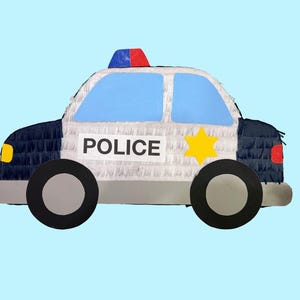 May include: A police car piñata with a blue, white, and black design. The piñata has a blue roof light, a yellow star, and the word "POLICE" on the side. The wheels are black and gray.