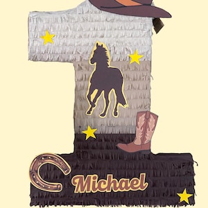 May include: A number one piñata with a cowboy theme. It features a brown cowboy hat, a horse silhouette, a cowboy boot, a horseshoe, and yellow star accents. The name "Michael" is written on the piñata.