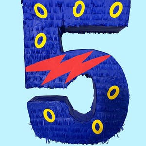 May include: A blue number five pinata featuring a textured fringe surface. The number is decorated with several yellow circular accents and a prominent red lightning bolt shape positioned across the center of the digit against a light blue background.