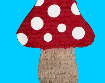 May include: A large, red mushroom-shaped piñata with white polka dots. The stem is beige and fringed. The piñata is set against a bright blue background.