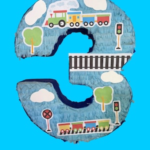 May include: A blue number three piñata decorated with train and railway-themed cutouts. The piñata features a green, yellow, and red train, white clouds, green trees, and a black and white railway track. A red and white railroad crossing sign and a traffic light are also included.