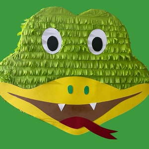 Peut inclure: Une grande piñata verte faite à la main en forme de tête de serpent de dessin animé. La piñata a de grands yeux blancs et noirs, un museau jaune, une bouche marron, des crocs blancs et une langue rouge. Le fond est vert uni.