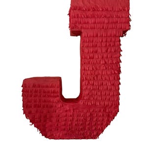 May include: A bright red letter J piñata covered in layers of fringed tissue paper. The structure is shaped like a capital letter J with a flat top and a curved base, featuring a textured, layered appearance created by the paper fringe.
