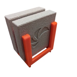 May include: A set of two gray, porous stone coasters with a carved design, held in an orange holder. The coasters are square and have a textured surface. The holder is a bright orange color and has a simple, geometric design.