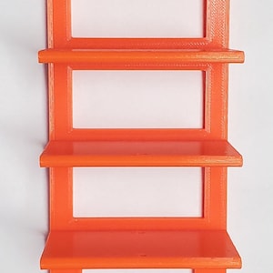 K&ouml;nnte beinhalten: Orangenes, 3D-gedrucktes Wandregal mit vier rechteckigen Ablagen. Das Regal hat eine rechteckige R&uuml;ckseite mit zwei L&ouml;chern zur Befestigung. Die Ablagen sind gleichm&auml;&szlig;ig angeordnet und haben eine Lippe an der Vorderkante. Das Regal ist aus Kunststoff.