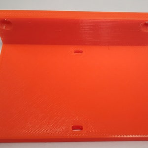 K&ouml;nnte beinhalten: Hellorangefarbene, 3D-gedruckte Kunststoffhalterung mit rechteckiger Basis und abgewinkeltem Oberteil. Das Oberteil hat zwei runde L&ouml;cher, die Basis zwei rechteckige Schlitze. Die Halterung ist f&uuml;r die Montage oder als St&uuml;tze konzipiert.
