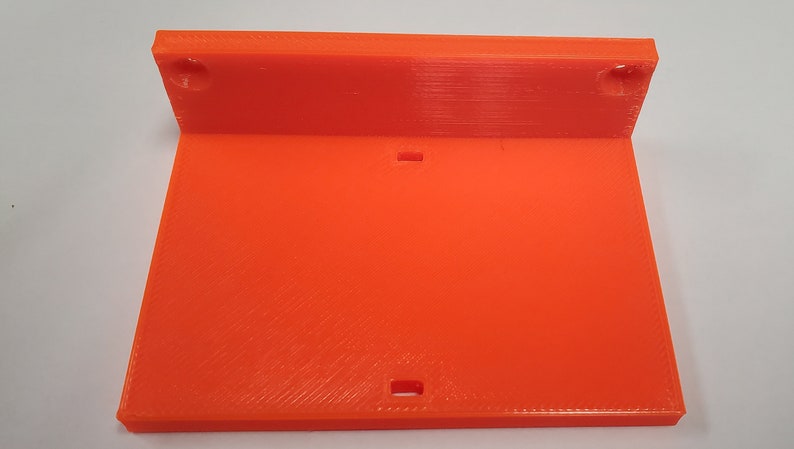 K&ouml;nnte beinhalten: Hellorangefarbene, 3D-gedruckte Kunststoffhalterung mit rechteckiger Basis und abgewinkeltem Oberteil. Das Oberteil hat zwei runde L&ouml;cher, die Basis zwei rechteckige Schlitze. Die Halterung ist f&uuml;r die Montage oder als St&uuml;tze konzipiert.