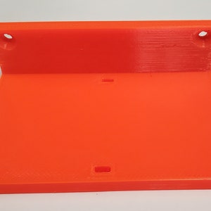 K&ouml;nnte beinhalten: Hellorangefarbene, 3D-gedruckte Regalhalterung. Die Halterung hat eine rechteckige Basis und ein R&uuml;ckenteil mit zwei kreisf&ouml;rmigen L&ouml;chern zur Befestigung. Auf der Basis befindet sich ein kleiner rechteckiger Ausschnitt. Die Halterung besteht aus Kunststoff.