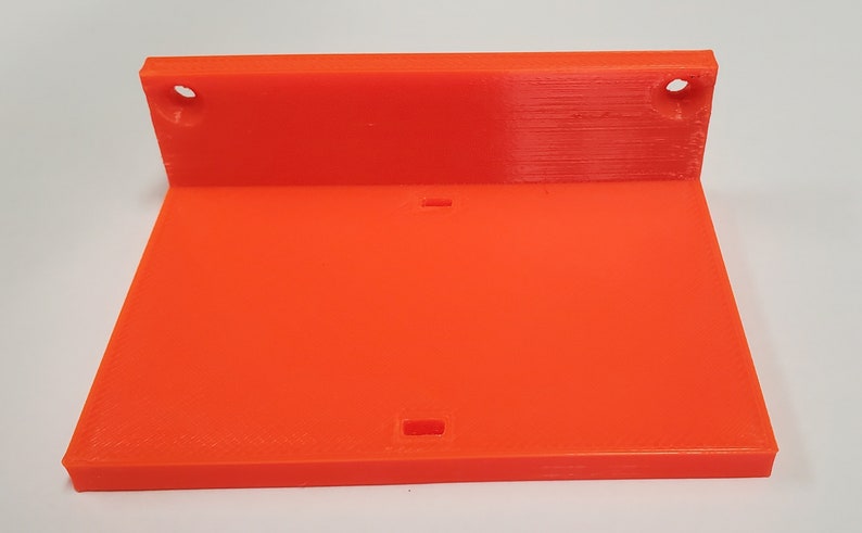 K&ouml;nnte beinhalten: Hellorangefarbene, 3D-gedruckte Regalhalterung. Die Halterung hat eine rechteckige Basis und ein R&uuml;ckenteil mit zwei kreisf&ouml;rmigen L&ouml;chern zur Befestigung. Auf der Basis befindet sich ein kleiner rechteckiger Ausschnitt. Die Halterung besteht aus Kunststoff.