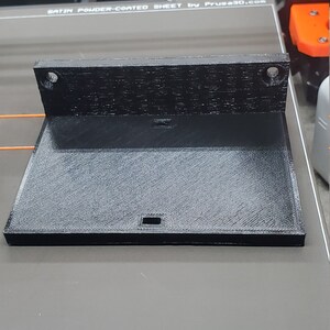 K&ouml;nnte beinhalten: Ein schwarzes 3D-gedrucktes Objekt, m&ouml;glicherweise ein Bauteil f&uuml;r einen 3D-Drucker, mit rechteckiger Basis und abgewinkeltem Oberteil. Das Objekt hat eine strukturierte Oberfl&auml;che und zwei L&ouml;cher oben. Die Basis hat einen kleinen rechteckigen Ausschnitt.