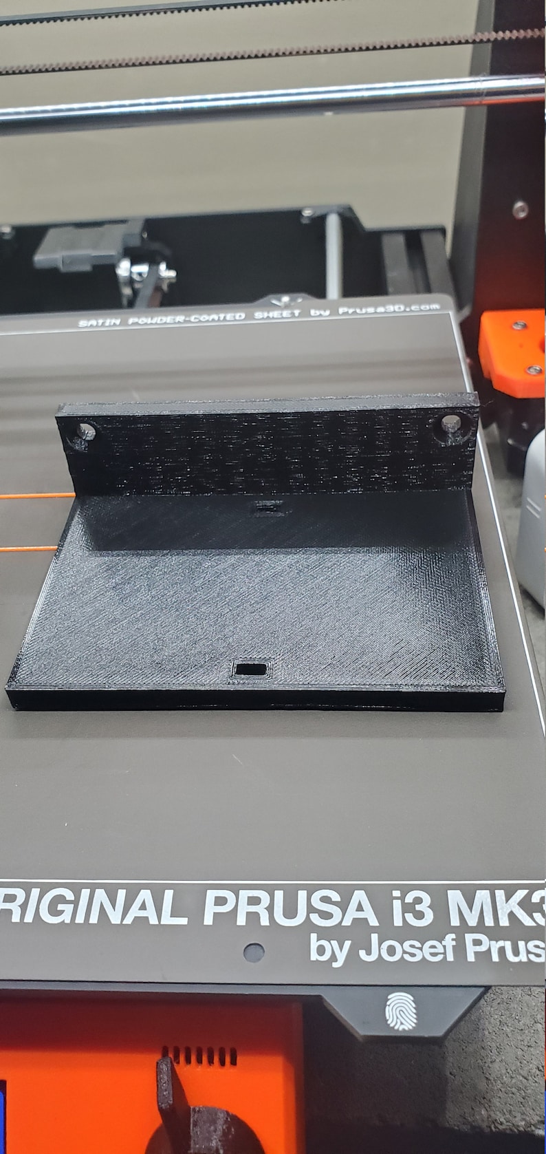 K&ouml;nnte beinhalten: Ein schwarzes 3D-gedrucktes Objekt, m&ouml;glicherweise ein Bauteil f&uuml;r einen 3D-Drucker, mit rechteckiger Basis und abgewinkeltem Oberteil. Das Objekt hat eine strukturierte Oberfl&auml;che und zwei L&ouml;cher oben. Die Basis hat einen kleinen rechteckigen Ausschnitt.