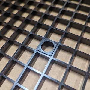 May include: Black plastic grid with a single square cutout in the center. The cutout has a round hole in the middle.
