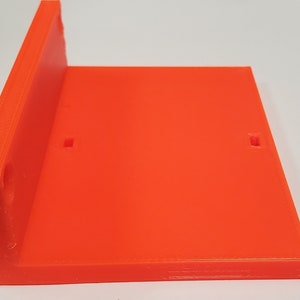 K&ouml;nnte beinhalten: Hellorangefarbene, rechtwinklige, 3D-gedruckte Kunststoffhalterung. Die Halterung hat eine flache Basis und eine senkrechte Seite mit zwei kleinen quadratischen Aussparungen an der Basis. Der Artikel dient zum Halten oder St&uuml;tzen anderer Objekte.