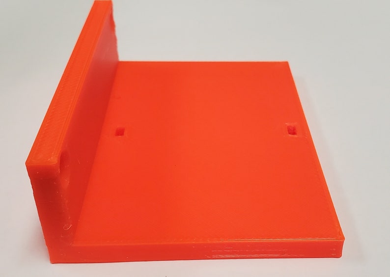 K&ouml;nnte beinhalten: Hellorangefarbene, rechtwinklige, 3D-gedruckte Kunststoffhalterung. Die Halterung hat eine flache Basis und eine senkrechte Seite mit zwei kleinen quadratischen Aussparungen an der Basis. Der Artikel dient zum Halten oder St&uuml;tzen anderer Objekte.