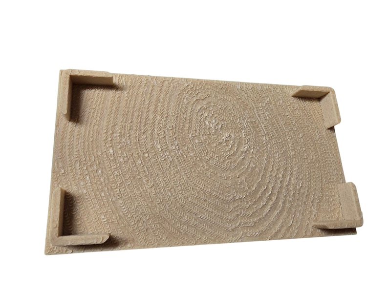 Pu&ograve; includere: Un vassoio rettangolare in legno marrone chiaro con una superficie strutturata che ricorda gli anelli degli alberi. Il vassoio ha quattro supporti angolari. Il design conferisce al vassoio un'estetica rustica e naturale.