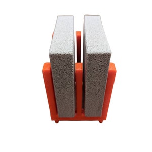 May include: A close-up of a shoe cleaner. The shoe cleaner has two gray rectangular blocks held in place by an orange plastic frame. The frame has a rectangular base and two vertical supports.