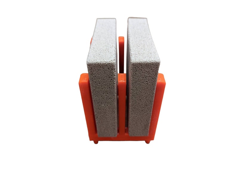 May include: A close-up of a shoe cleaner. The shoe cleaner has two gray rectangular blocks held in place by an orange plastic frame. The frame has a rectangular base and two vertical supports.