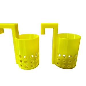 May include: Two bright yellow, 3D-printed, over-the-door cup holders. Each holder has a cylindrical cup with circular cutouts and a rectangular hook for hanging. The design is simple and functional.