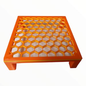 May include: Orange and yellow honeycomb-patterned plastic stand with a raised base. The stand is made of 3D printed plastic.