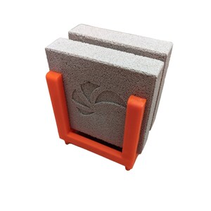 May include: A set of three gray, porous stone coasters with a floral design, held in an orange plastic holder. The coasters are stacked vertically, showcasing their textured surface. The holder provides a vibrant contrast.