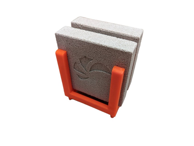 May include: A set of three gray, porous stone coasters with a floral design, held in an orange plastic holder. The coasters are stacked vertically, showcasing their textured surface. The holder provides a vibrant contrast.