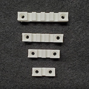 May include: Four white plastic mounting brackets of varying sizes are arranged vertically. Each bracket has a series of ridges and holes for screws. The brackets are on a dark gray textured surface.