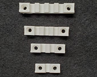 May include: Four white plastic mounting brackets of varying sizes are arranged vertically. Each bracket has a series of ridges and holes for screws. The brackets are on a dark gray textured surface.