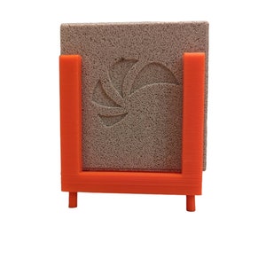May include: An orange, 3D-printed holder with a square, textured, gray stone. The stone has a floral design carved into it. The holder has a U-shape with two legs for support.
