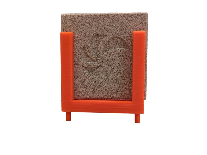 May include: An orange, 3D-printed holder with a square, textured, gray stone. The stone has a floral design carved into it. The holder has a U-shape with two legs for support.