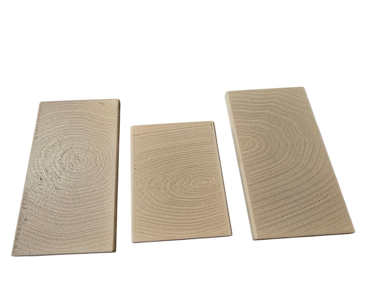 Pu&ograve; includere: Tre blocchi di legno non rifiniti con motivi di venature visibili. I blocchi sono rettangolari, uno dei quali ha una forma trapezoidale. Il legno ha un colore chiaro e naturale, e la venatura crea motivi circolari.