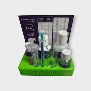 May include: A green plastic tray holds a calcium test kit. The kit includes syringes, vials, a clear glass, and a reference solution bottle. A purple card with the text "Aquaforest TestPro" and a calcium concentration chart is in the background.
