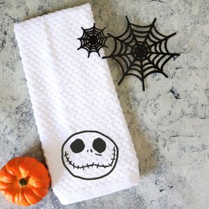 Może przedstawiać: Biały ręcznik kuchenny o tematyce Halloween z motywem twarzy Jacka Skellingtona. Ręcznik jest złożony i ma wzorzystą fakturę. Obecne są r&oacute;wnież dwie czarne dekoracje z pajęczyny i mała pomarańczowa dynia.