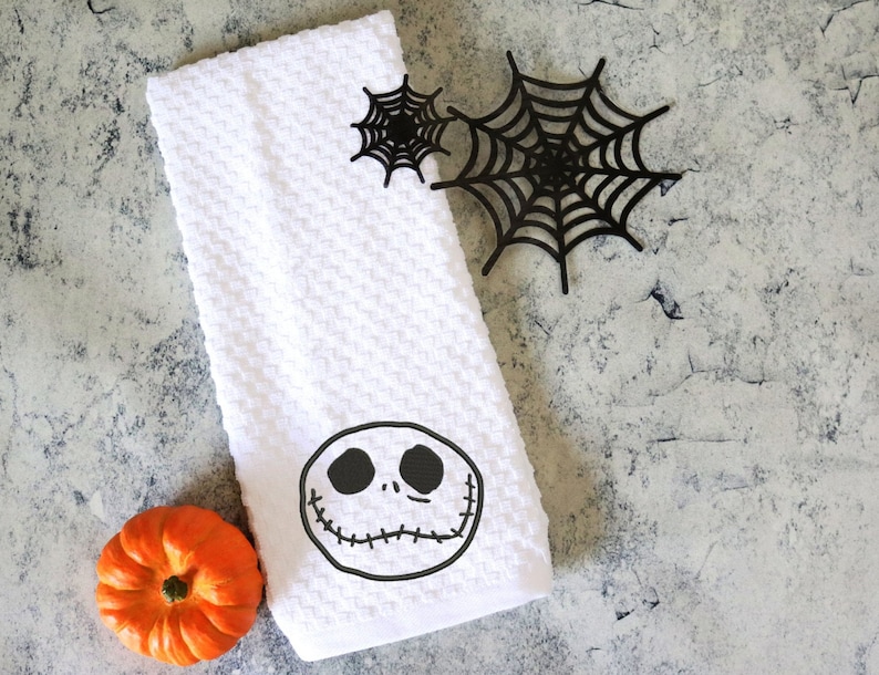 Może przedstawiać: Biały ręcznik kuchenny o tematyce Halloween z motywem twarzy Jacka Skellingtona. Ręcznik jest złożony i ma wzorzystą fakturę. Obecne są r&oacute;wnież dwie czarne dekoracje z pajęczyny i mała pomarańczowa dynia.
