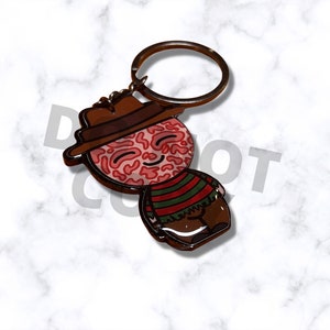May include: A novelty keychain featuring a cartoon character with a brown hat, a red and brown face, and a striped sweater. The keychain has a bronze-colored ring for attaching to keys or a bag. The character's face has a smiling expression.