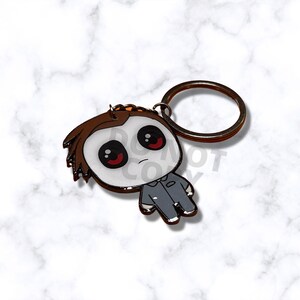 May include: A cartoon-style keychain featuring a character with a white mask, brown hair, and a gray jumpsuit. The character has large black eyes with red pupils. The keychain has a bronze-colored ring.
